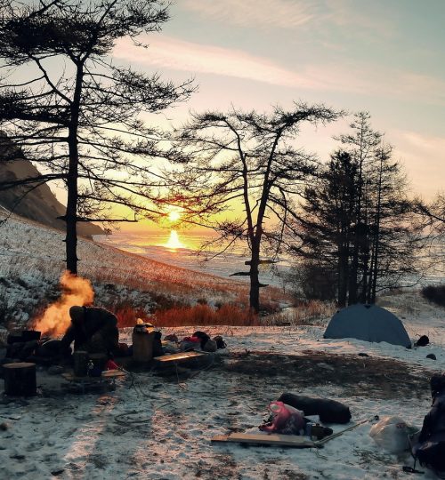 Winter camping scene with bonfire and bare trees at sunset in snow